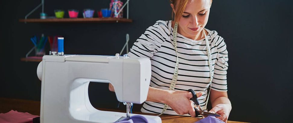 Young woman in blue and white stripped shirt cutting fabric while sitting behind a sewing machine